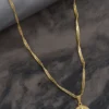 Un collar de cadena de oro con un colgante redondo que presenta una figura grabada descansa sobre una superficie oscura y texturizada junto a una tela enrollada de color gris. Esta elegante pieza es parte de nuestra exquisita colección de Joyas en Oro Laminado 18K.