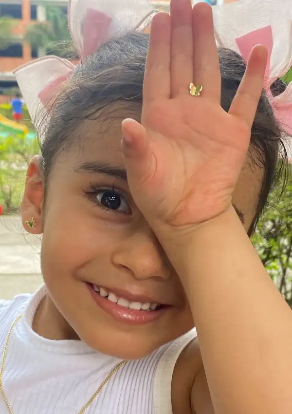 Un niño pequeño posa al aire libre con una gran sonrisa, tapándose un ojo con la mano. El niño lleva pendientes de oro Arete Bright Star y un collar a juego, con lazos rosas en el pelo.