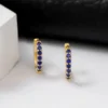 Pendientes de aro de oro con piedras preciosas azules expuestas sobre una superficie blanca sobre un fondo negro.