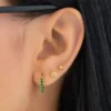 Primer plano de la oreja de una persona con tres piercings: un aro verde, un pendiente redondo con una piedra transparente y un pendiente pequeño de oro. Se ven el pelo negro y la montura de las gafas.