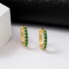 Par de pequeños pendientes de aro dorados adornados con piedras preciosas verdes, colocados sobre una superficie blanca con cuero negro de fondo.