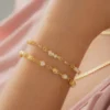 Pulsera de cadena de oro adornada con pequeñas perlas que se lleva en la muñeca, sobre un fondo de tela de color rosa claro.