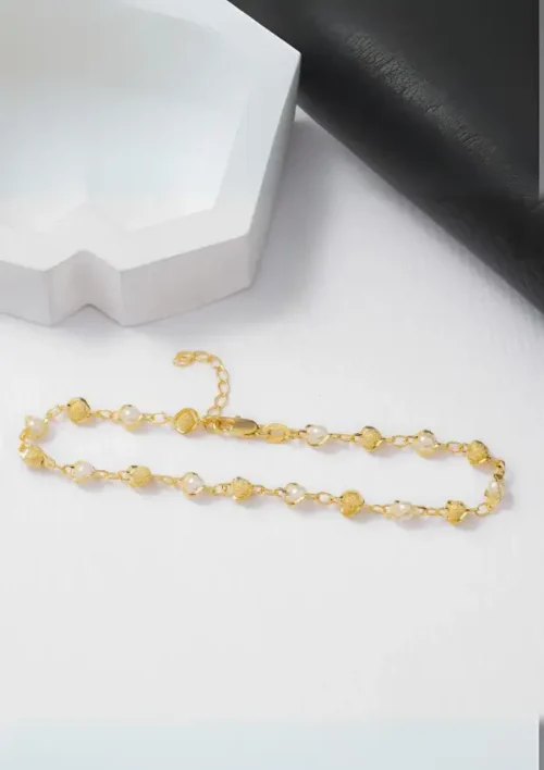 Pulsera de oro y perlas con cadena ajustable expuesta sobre una superficie blanca junto a contenedores con formas geométricas.