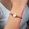 Un primer plano de la Pulsera San Benito con Rondel, que presenta cuentas doradas y rojas con un dije de moneda, elegantemente exhibida en la muñeca de una persona.