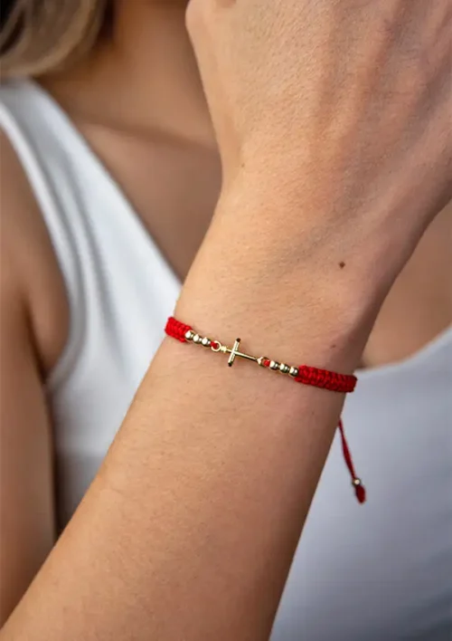 Una persona que lleva en su muñeca la "Pulsera con Herraje de Cruz Lisa", una pulsera tejida de color rojo con un diseño de cruz horizontal.