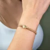 La "Pulsera San Benito Tradicional herraje pequeño", una pulsera de oro con un pequeño dije redondo, se usa en la muñeca de una persona, exhibida sobre una parte superior blanca.