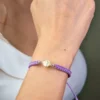 Una persona que lleva la Pulsera San Benito Tradicional herraje pequeño, que presenta un diseño trenzado de color morado con un dije redondo de color dorado en su muñeca.