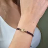 Una persona que lleva la Pulsera San Benito Tradicional herraje pequeño, una pulsera trenzada marrón con un dije redondo de oro en su muñeca.