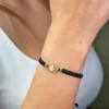 Una persona que lleva la Pulsera San Benito Tradicional herraje pequeño en su muñeca, que presenta un diseño tejido en negro con un pequeño dije redondo dorado.