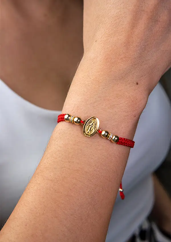 Una persona que lleva en su muñeca la Pulsera con Herraje de la Virgen Milagrosa con Rondel, que consta de un cordón rojo con cuentas de oro y un medallón de oro.