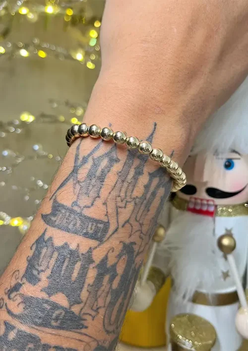 Se muestra un brazo tatuado que lleva la Pulsera Full Balín lisa dorada de 0,7 milímetros, con un cascanueces decorativo en el fondo.