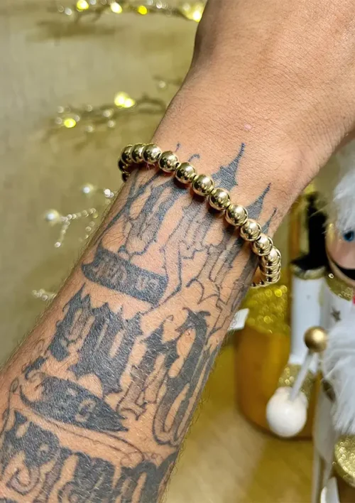 Un antebrazo tatuado adornado con la Pulsera Full Balín lisa dorada de 0,8 milímetros, sobre un fondo borroso acentuado por luces de cadena y una figura decorativa blanca.