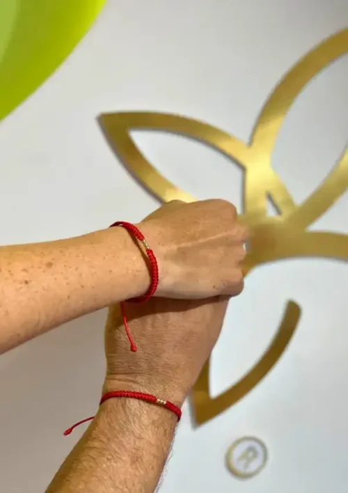 Dos manos adornadas con Pulseras 3 balines con brazaletes de hilo rojo se mantienen juntas frente a una pared que muestra un diseño floral abstracto dorado.