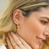 Una mujer de cabello rubio luce los pendientes y anillos de oro Aretes Candongas Inspire 3cms, con una mano tocando suavemente su cuello.