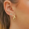 Oreja de mujer adornada con tres pendientes de oro: un pequeño botón, un botón con forma de diamante y el aro mediano Aetes Twisted Stripes de 2 cm.