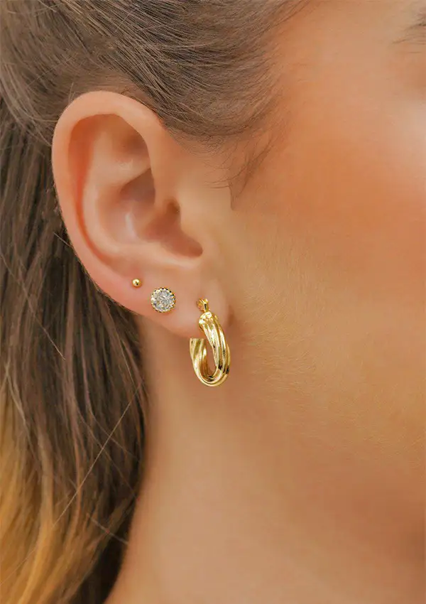 Oreja de mujer adornada con tres pendientes de oro: un pequeño botón, un botón con forma de diamante y el aro mediano Aetes Twisted Stripes de 2 cm.