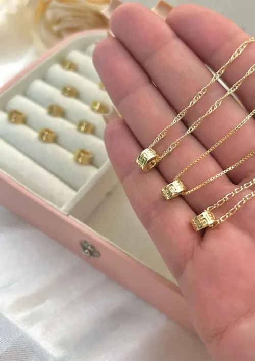 En una mano se encuentran tres collares de oro “Herraje medusa” con pequeñas letras colgantes, acompañados de un joyero abierto con anillos de oro adicionales.