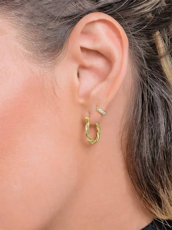 Primer plano de la oreja de una mujer adornada con un aro de oro Candonga mini C. de 8 mm y un pendiente de puño de oro con pequeñas piedras en la parte superior de la oreja; el cabello está recogido.