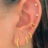 Primer plano de una oreja que muestra múltiples perforaciones: dos aretes tipo botón, un arete en forma de corazón y tres aros dorados CANDONGA ESFERA P con detalle de bolas.