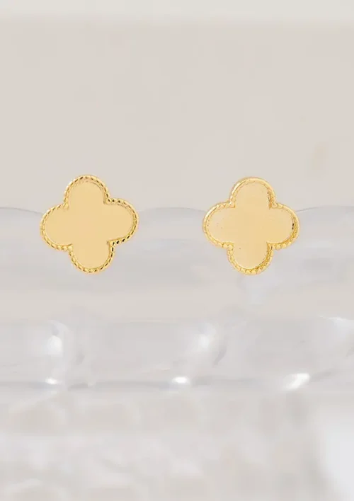 Los pendientes Arete clover cms presentan un diseño dorado en forma de trébol con un borde texturizado colocado sobre una superficie clara.