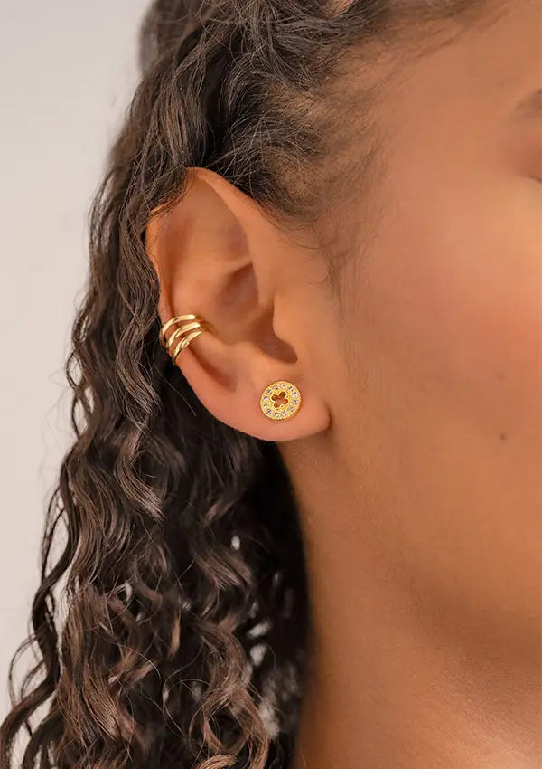 Un primer plano muestra una oreja adornada con los Aretes Clover Redondo silueta 9mm, que presenta pequeños aretes circulares con piedras rojas y amarillas, junto a tres ear cuffs dorados en la parte superior de la oreja.