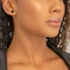 Una persona con cabello rizado lleva el "Conjunto Aretes y dije Clover Negro 1cm", que incluye un collar en forma de trébol y aretes a juego. Muestra una expresión neutra y usa un top oscuro.