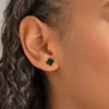 Primer plano de alguien que lleva el "Conjunto Aretes y dije Clover Negro 1cm", que presenta un botón negro en forma de trébol con bordes dorados.