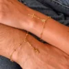 Dos manos adornadas con delicadas pulseras de cadena de oro Denario 17 CMS con dijes de cruces, que reposan sobre tela vaquera.