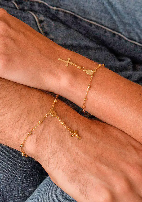 Dos manos adornadas con delicadas pulseras de cadena de oro Denario 17 CMS con dijes de cruces, que reposan sobre tela vaquera.