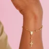 La pulsera de oro Denario 18,5 CMS con una pequeña cruz cuelga de la muñeca de una persona sobre un fondo rosa.