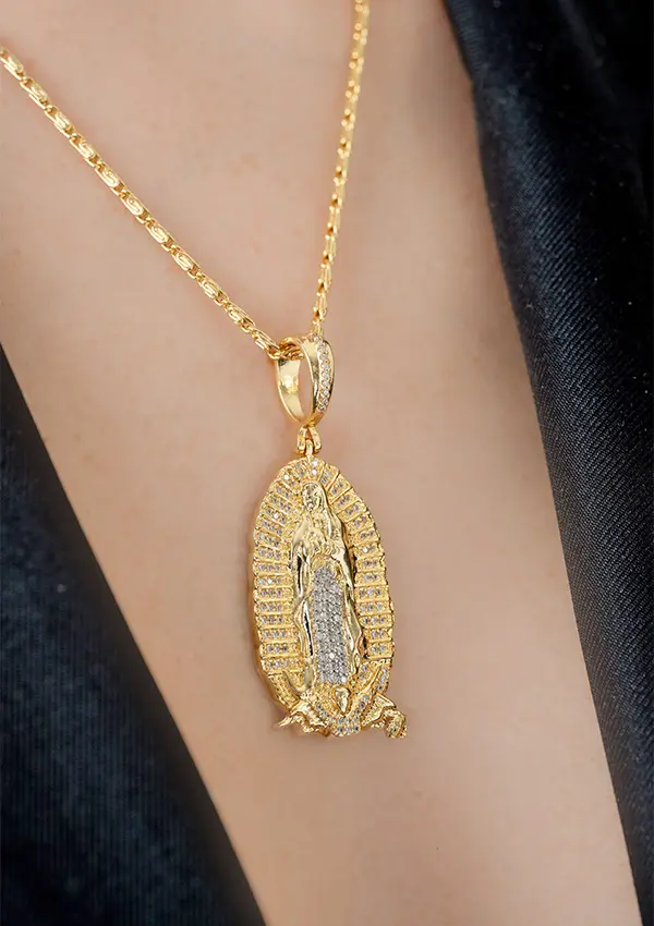 El Dije Nuestra Señora de Guadalupe circonias es un collar de oro con un colgante ovalado grabado con una figura, que se lleva elegantemente sobre tela oscura.