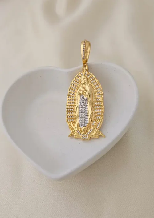 Dije Nuestra Señora de Guadalupe en oro y plata con circonitas, expuesto en plato blanco en forma de corazón sobre fondo claro.