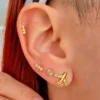 Primer plano de una oreja adornada con unos pendientes de oro con circones de Arete Avión, que luce tachuelas y un diseño floral, mientras una mano con esmalte de uñas rosa toca delicadamente la oreja.