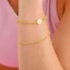 Una persona lleva dos pulseras de oro, incluida la "Pulsera Militar 1.1 mm de 18cms", con pequeñas cuentas y un dije circular, sobre un fondo rosa borroso.