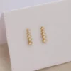 Aretes Guadalupe Finita son unos aretes de oro que presentan múltiples pequeñas piedras tipo diamante, dispuestas verticalmente sobre una superficie blanca.