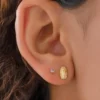 Primer plano de una oreja adornada con dos aretes: un pequeño stud redondo y los Aretes Guadalupe Finita, un arete ovalado de oro que presenta un diseño detallado.