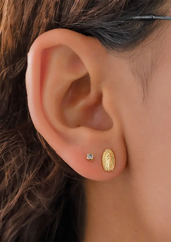 Primer plano de una oreja adornada con dos aretes: un pequeño stud redondo y los Aretes Guadalupe Finita, un arete ovalado de oro que presenta un diseño detallado.